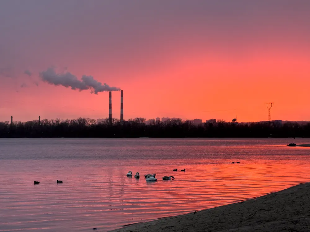 Sunset over the Dnipro River in Kyiv with swans on the water and smoking power plant chimneys.